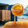 Envío de miel premium para exportación, dos contenedores llenos de cajas de miel en un moderno puerto marítimo internacional, tarro de miel dorado con miel fluyendo y panal natural en primer plano, carretilla elevadora cargando palés de exportación, buque de carga al fondo, sutil concepto comercial de Malasia, fotografía profesional de exportación de alimentos B2B, ultrarrealista, cálida luz solar dorada, logística global, comercio internacional, estilo publicitario comercial, muy detallado, enfoque nítido, marca premium, relación de aspecto 16:9, calidad 8K, sin texto, sin marca de agua